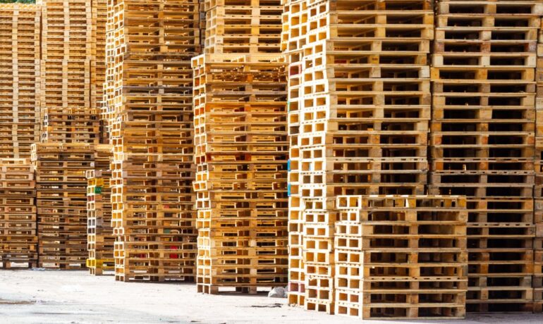 Wooden pallets are stacked up to varying heights on a concrete surface outdoors on a sunny day near trees.