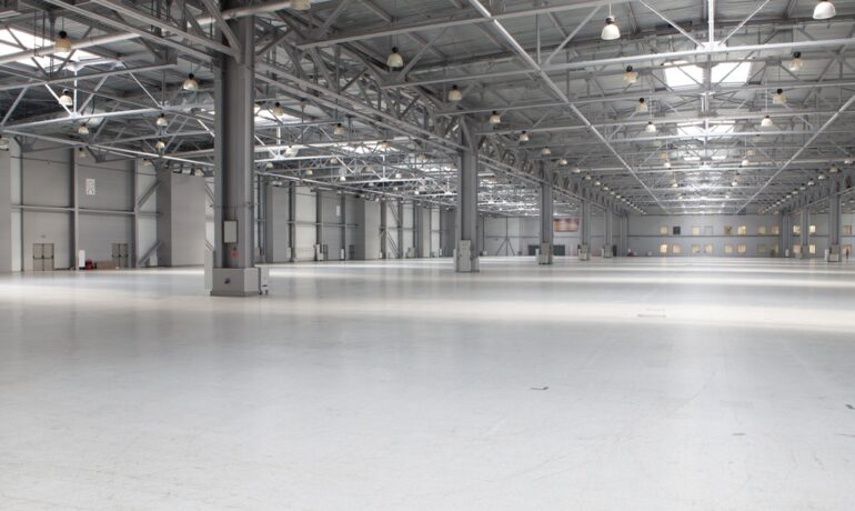 A large, empty, gray and white modern warehouse with exposed steel beams and rows of hanging light fixtures and skylights.