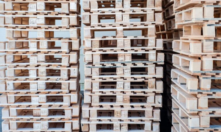 Wooden pallets are stacked in multiple tall columns while sitting on a well-lit warehouse floor near a white wall.