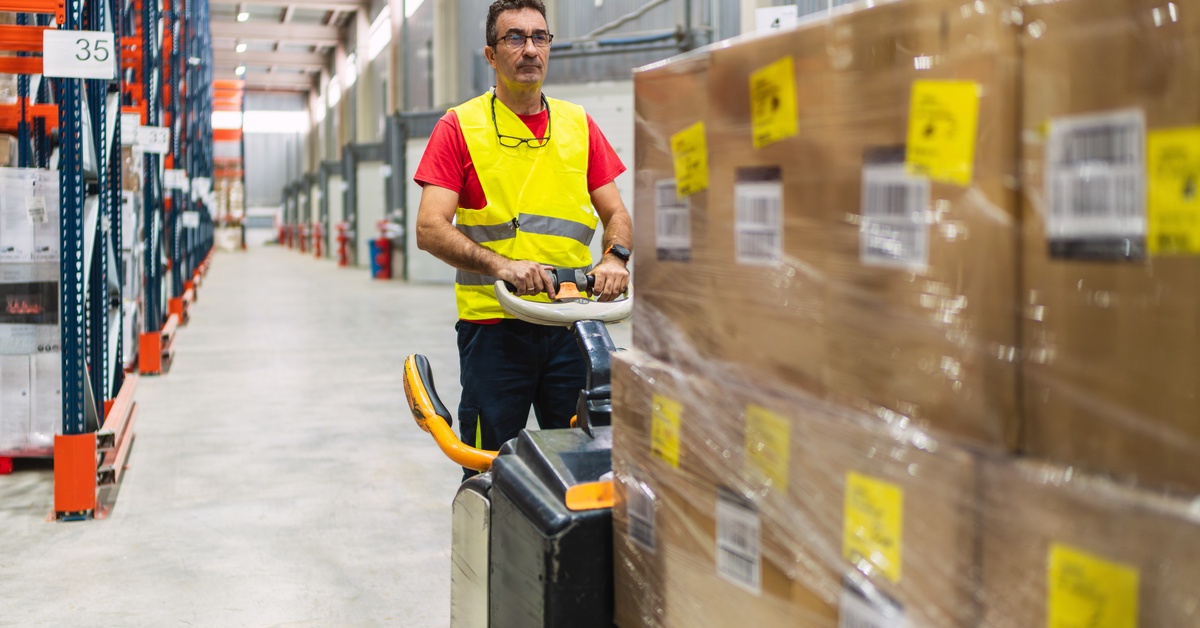 A warehouse worker drives a forklift stacked with boxes across a large facility with shelving lined up in a row on one side.