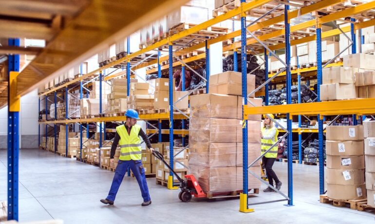 falm-3643Two warehouse workers wearing hard hats use a pallet jack to move boxes in a clean, organized warehouse.