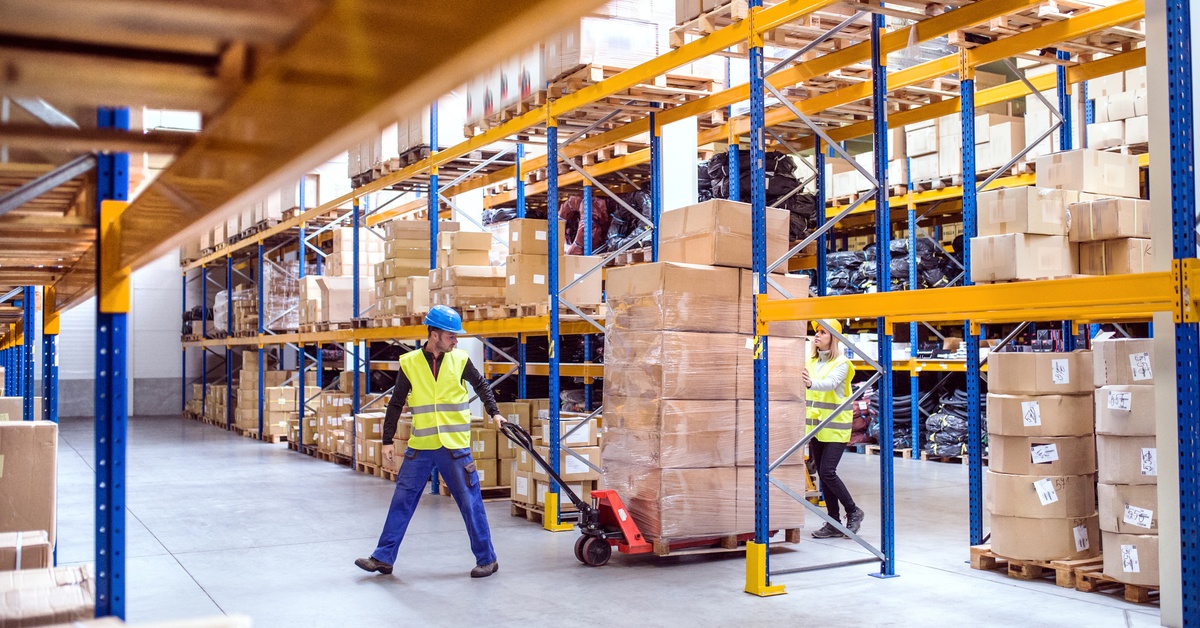 falm-3643Two warehouse workers wearing hard hats use a pallet jack to move boxes in a clean, organized warehouse.