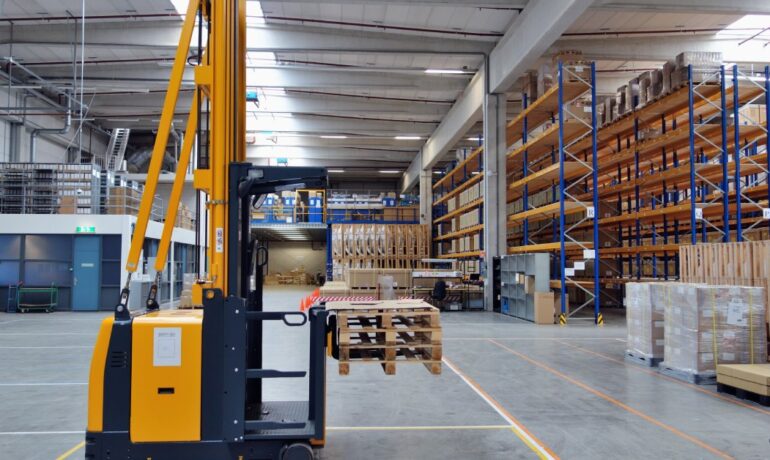 A forklift holds some pallets in an open space in a warehouse with blue shelving that extends to the ceiling.