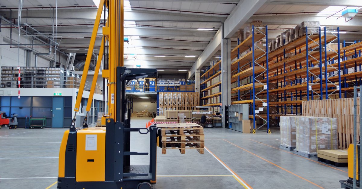A forklift holds some pallets in an open space in a warehouse with blue shelving that extends to the ceiling.