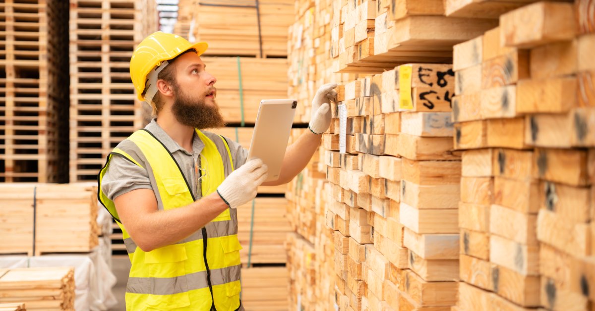 A man wearing a hard hat, high-vis vest, and white gloves holds a tablet while checking stacks of wood.