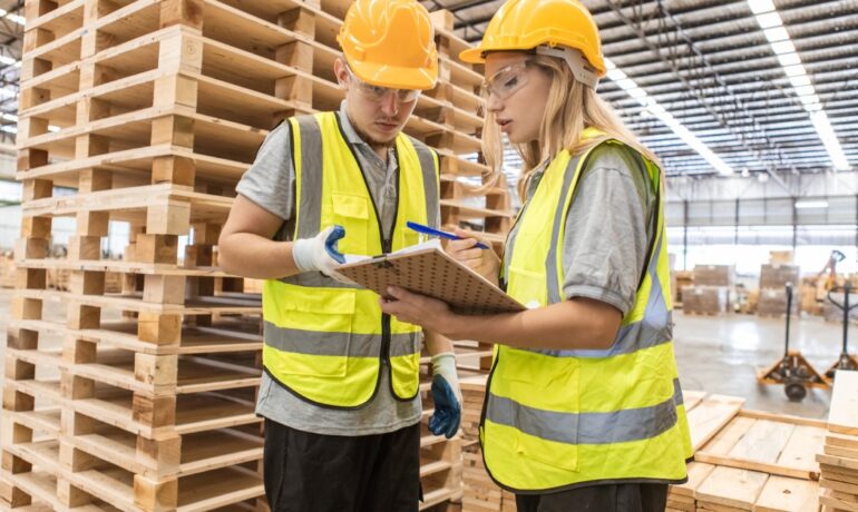 Two workers in protective gear review paperwork while standing in front of a tall stack of wooden pallets.