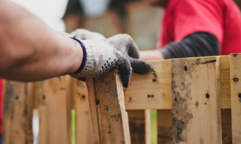 A person wearing gloves removes a wooden slat from a pallet for repair and recycling while another person assists nearby.