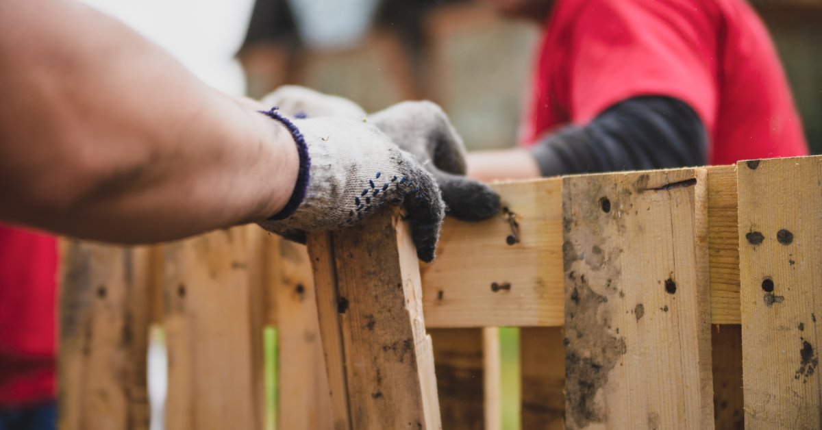 A person wearing gloves removes a wooden slat from a pallet for repair and recycling while another person assists nearby.