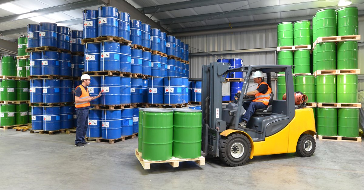 Inside a metal warehouse, two men use a forklift to move green and blue barrels of chemicals on pallets.