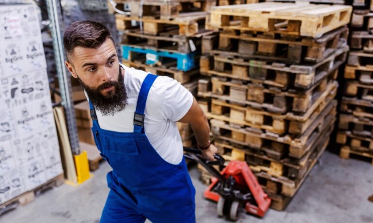 A man in coveralls uses a pallet jack to move wood pallets in a warehouse. Stacked pallets are behind him.