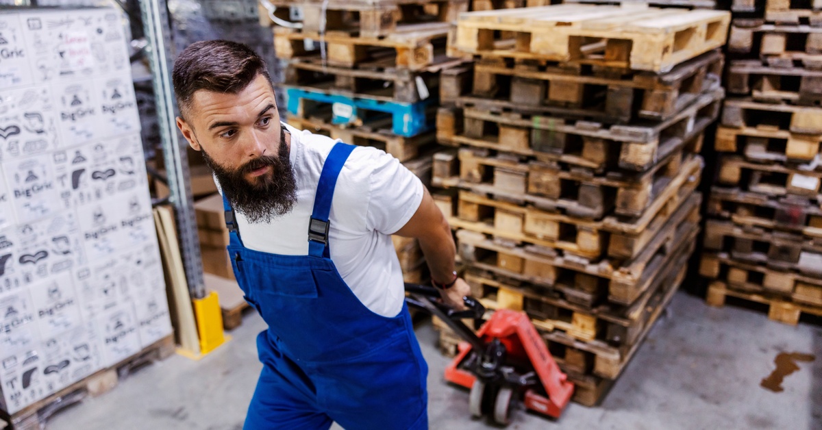 A man in coveralls uses a pallet jack to move wood pallets in a warehouse. Stacked pallets are behind him.