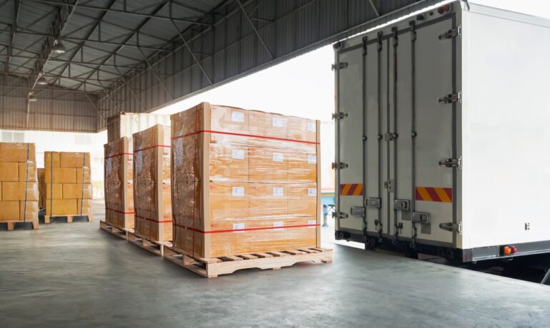 Boxes are stacked and wrapped on a wooden pallet near a cargo truck backed up to a loading dock with its doors closed.