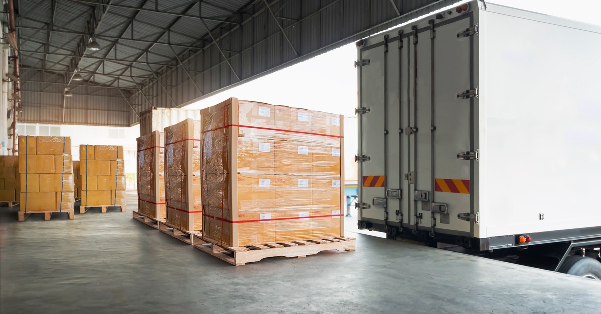 Boxes are stacked and wrapped on a wooden pallet near a cargo truck backed up to a loading dock with its doors closed.