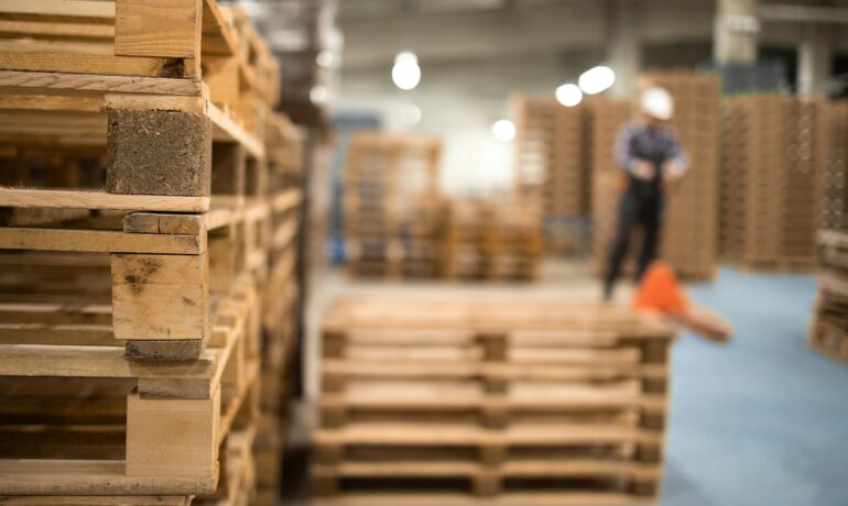 A stack of wooden pallets is in focus inside a warehouse, with a blurred worker wearing a hard hat visible in the background.