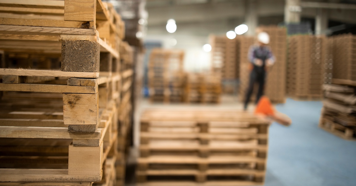 A stack of wooden pallets is in focus inside a warehouse, with a blurred worker wearing a hard hat visible in the background.