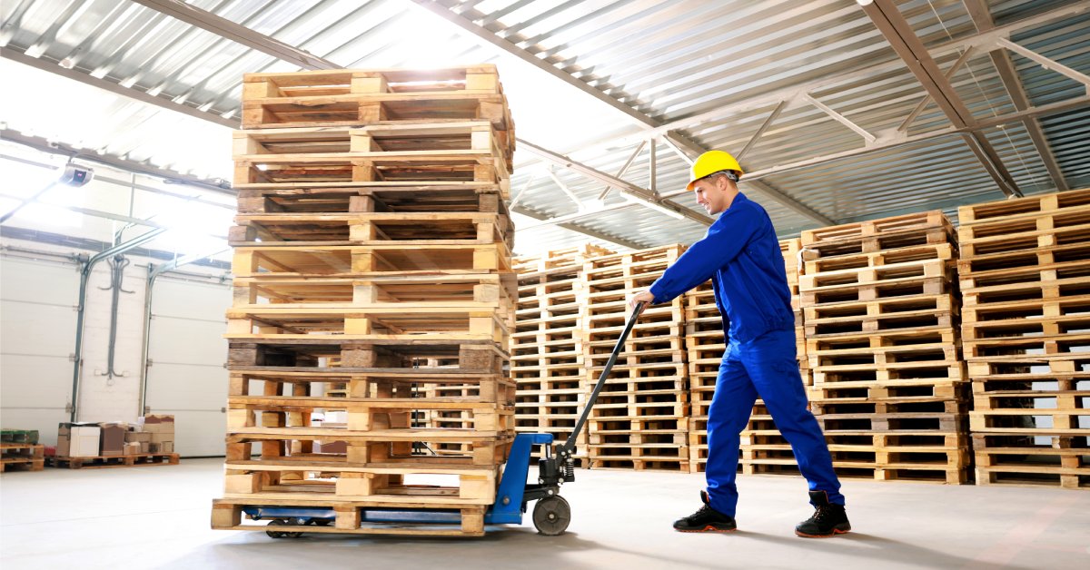 A worker uses a pallet jack to move wood pallets in a warehouse. Stacked pallets are behind him.