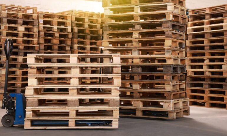 A blue pallet jack is positioned under wooden pallets beside several taller pallet stacks inside a warehouse.