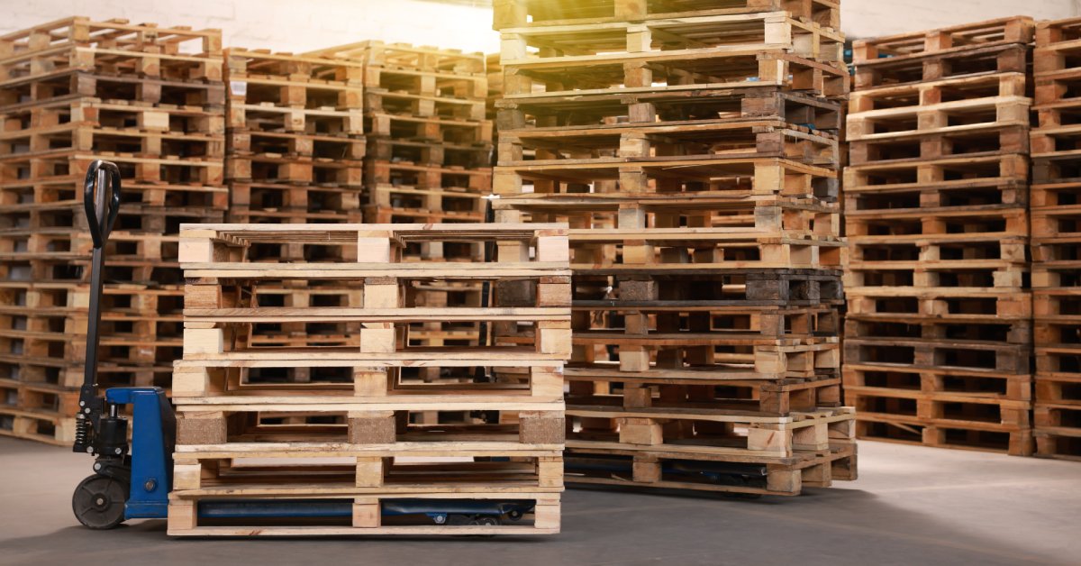 A blue pallet jack is positioned under wooden pallets beside several taller pallet stacks inside a warehouse.