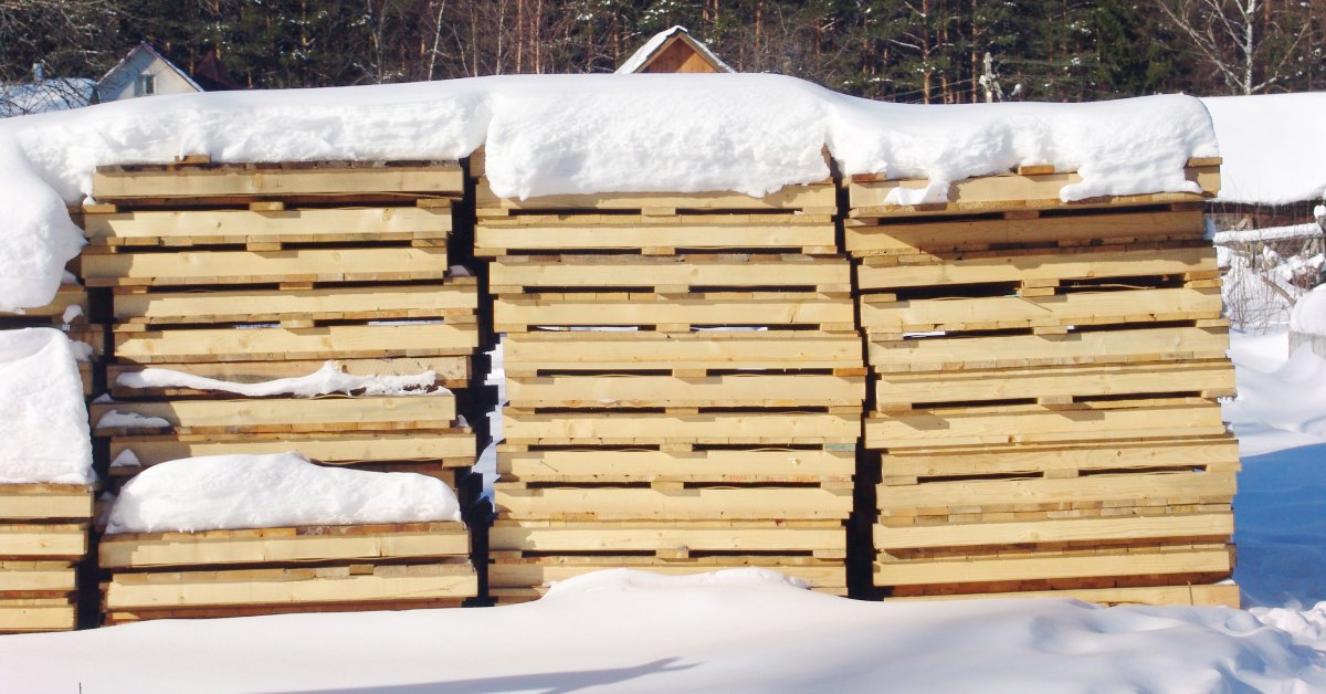 Stacks of wooden pallets sit side by side outdoors, with snow covering the tops and the ground around them.