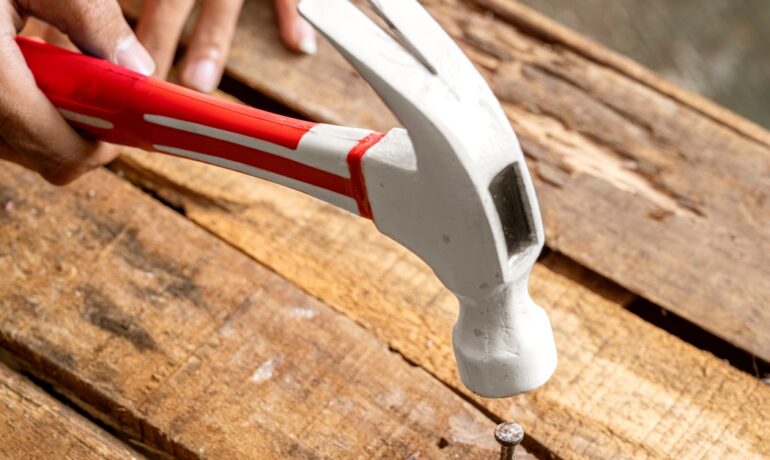 A person uses a hammer with a red handle to nail down a wooden board that is popping up from a pallet.