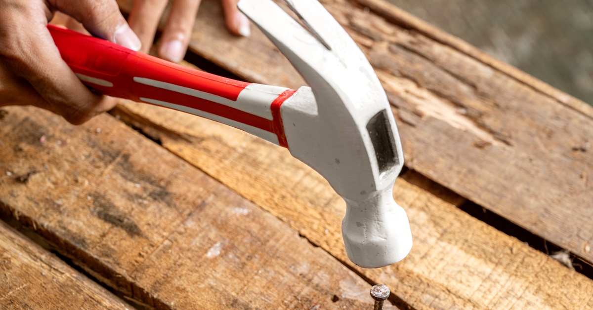 A person uses a hammer with a red handle to nail down a wooden board that is popping up from a pallet.