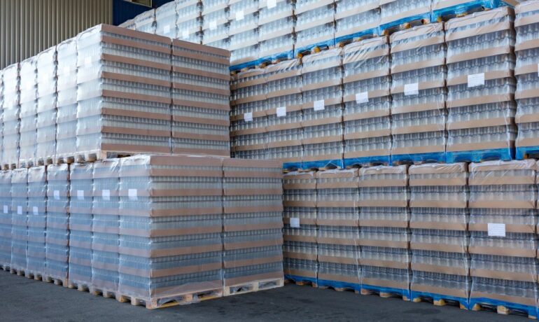 Clear glass bottles are packed in cardboard, wrapped in plastic, and stacked on wooden pallets in open-air storage.