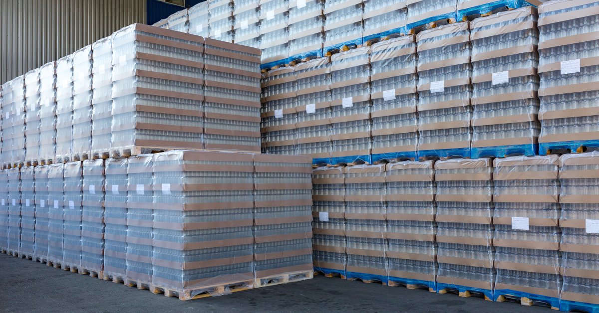 Clear glass bottles are packed in cardboard, wrapped in plastic, and stacked on wooden pallets in open-air storage.