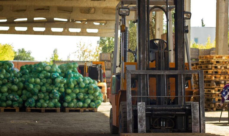 An empty forklift is parked outdoors near stacks of food on wood pallets under a covered factory area.