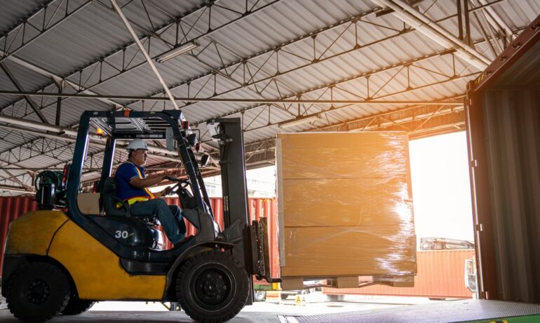 A warehouse worker drives a forklift carrying a boxed pallet and loads it into a large shipping container for transport.