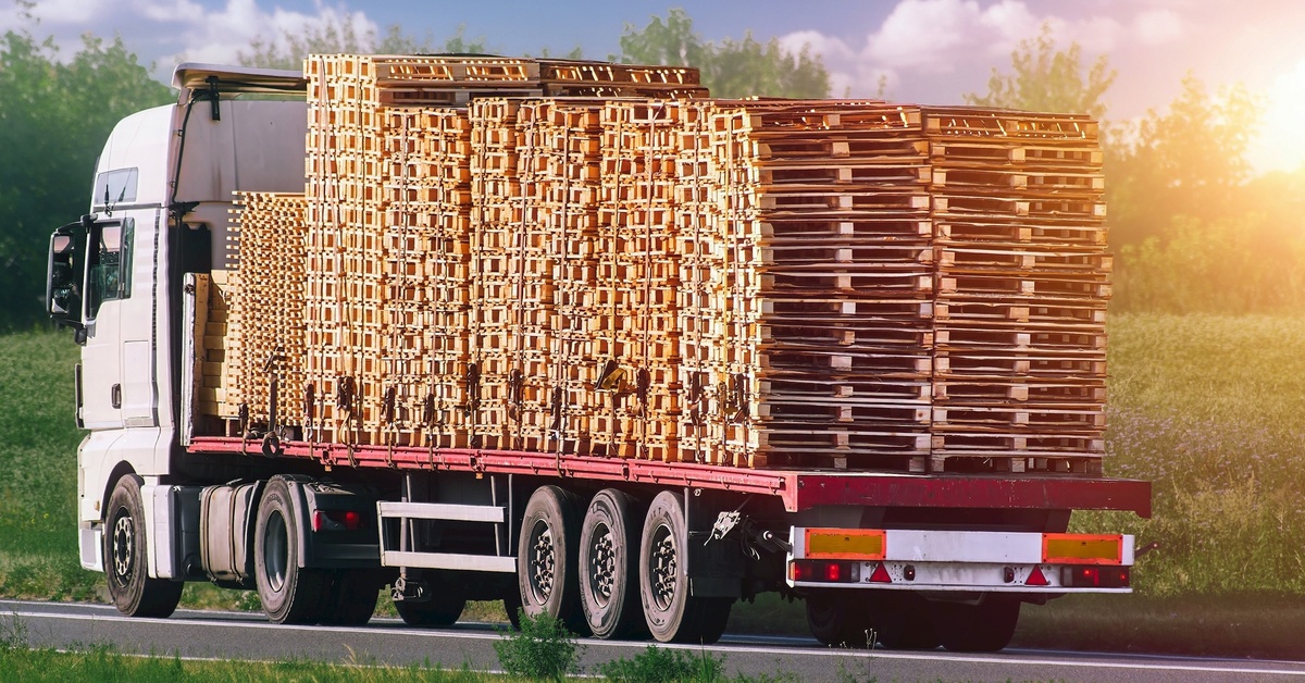 A flatbed truck loaded up with hundreds of wooden pallets, stacked in two rows and strapped to the bed, down a country road.