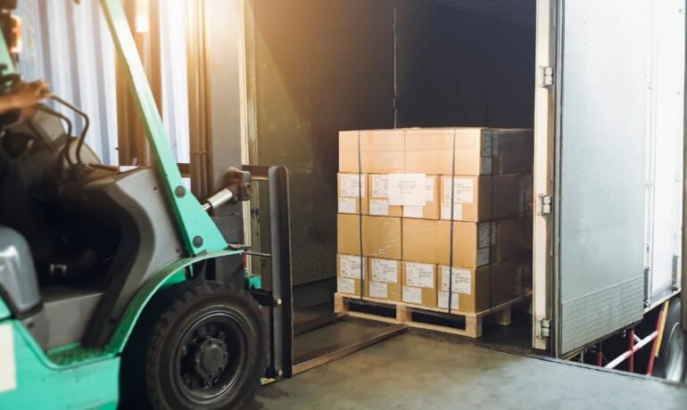 A forklift loads stacked boxes on a pallet into a truck trailer at a busy warehouse loading dock area.