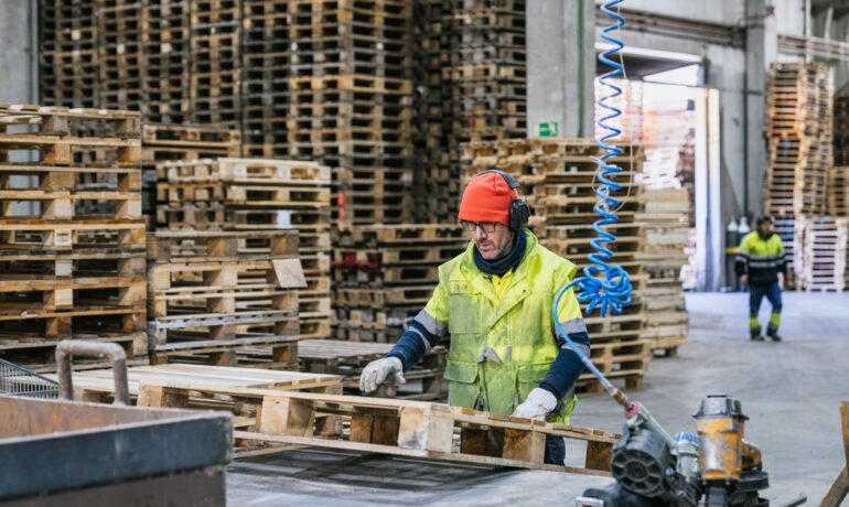 Worker in safety gear handling wooden pallet in warehouse with stacked pallets and industrial equipment.