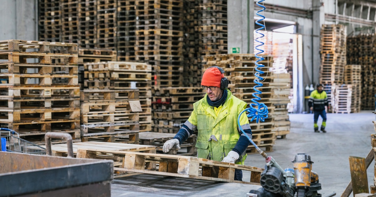 Worker in safety gear handling wooden pallet in warehouse with stacked pallets and industrial equipment.