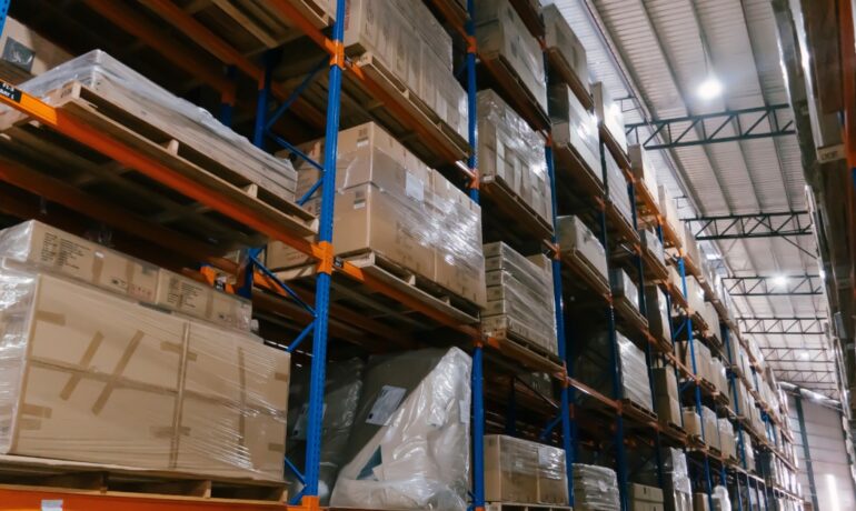 Tall warehouse shelving stocked with boxed inventory and palletized goods in a large distribution center.