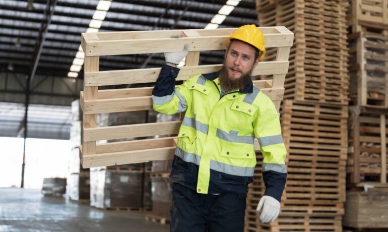 A warehouse worker in a safety jacket and helmet carries a wooden pallet on his shoulder inside a storage facility.