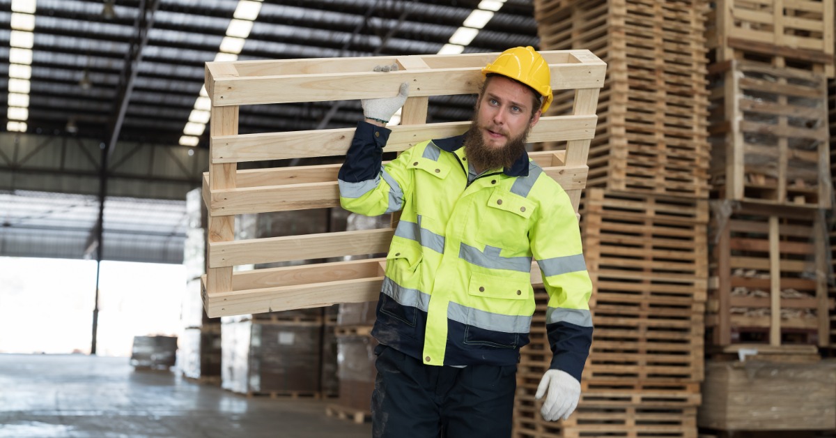 A warehouse worker in a safety jacket and helmet carries a wooden pallet on his shoulder inside a storage facility.