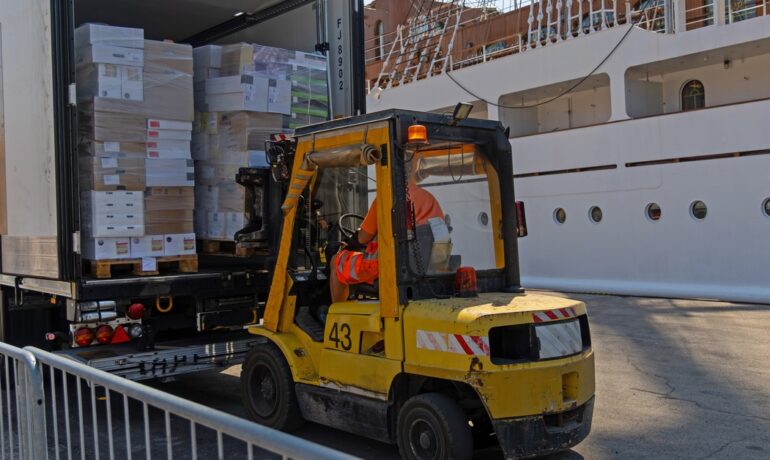A forklift loads palletized goods into a truck near a docked cargo ship, showing active shipping operations.