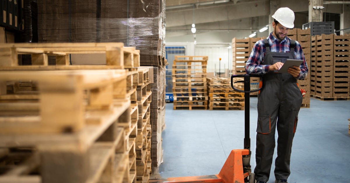 A warehouse worker uses a tablet beside a pallet jack while inspecting stacks of wooden pallets in a storage area.