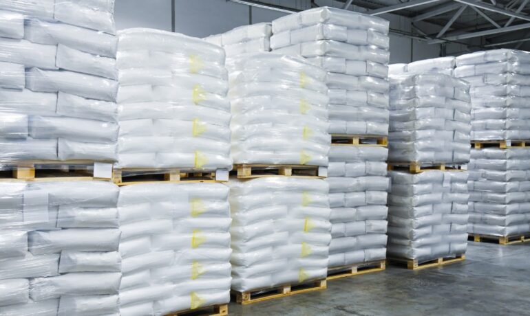 Wooden pallets loaded with white bagged products are stacked two high in rows inside an industrial warehouse.