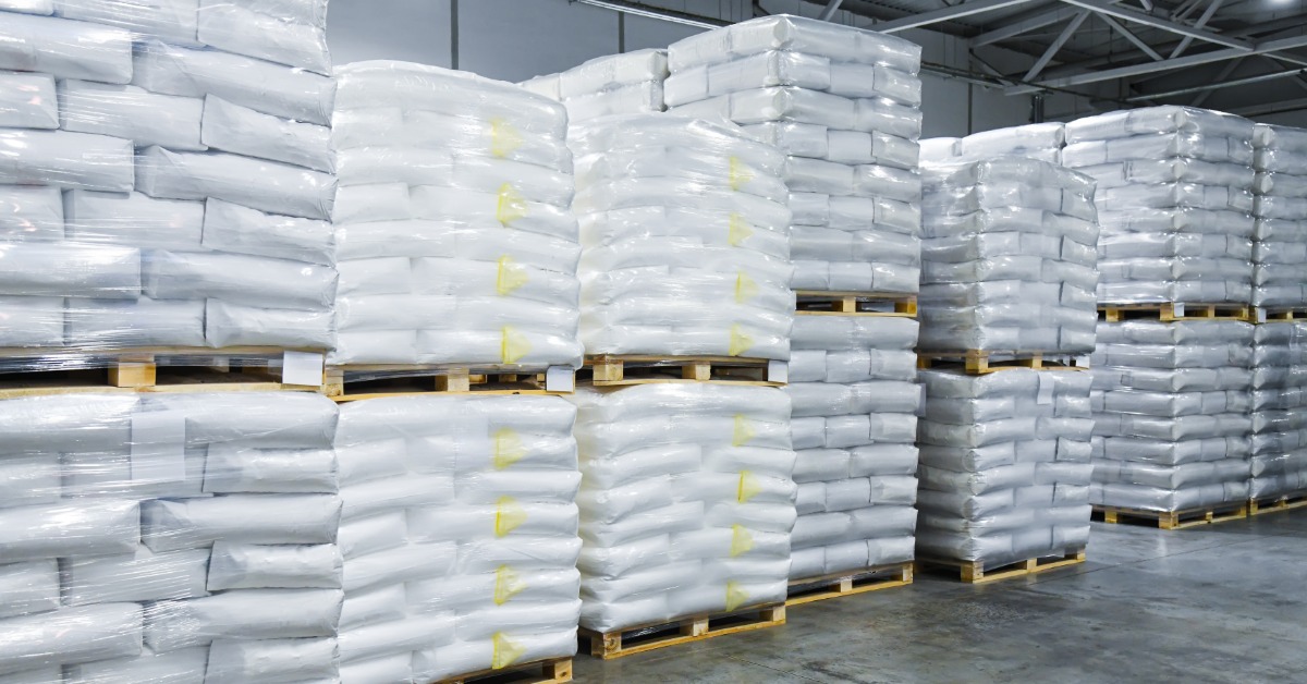 Wooden pallets loaded with white bagged products are stacked two high in rows inside an industrial warehouse.