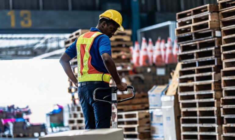 A warehouse worker operates a pallet jack beside stacks of wooden pallets staged inside a distribution facility.