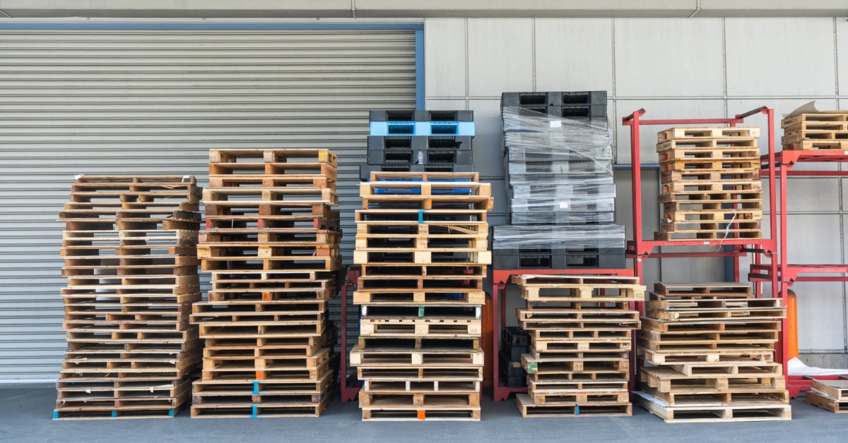 Stacks of wooden pallets are organized inside a warehouse with a pallet jack positioned among the available inventory.