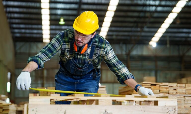 A warehouse worker wearing a yellow hard hat measures a wooden pallet with a tape measure to check its dimensions.