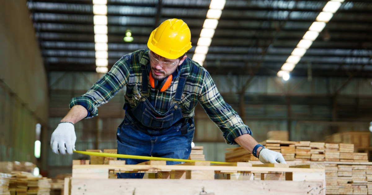 A warehouse worker wearing a yellow hard hat measures a wooden pallet with a tape measure to check its dimensions.